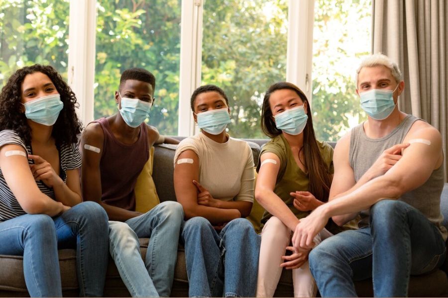 A group of young people wearing PPE face masks over their mouths and noses with plasters on their upper arms where they've had an injection