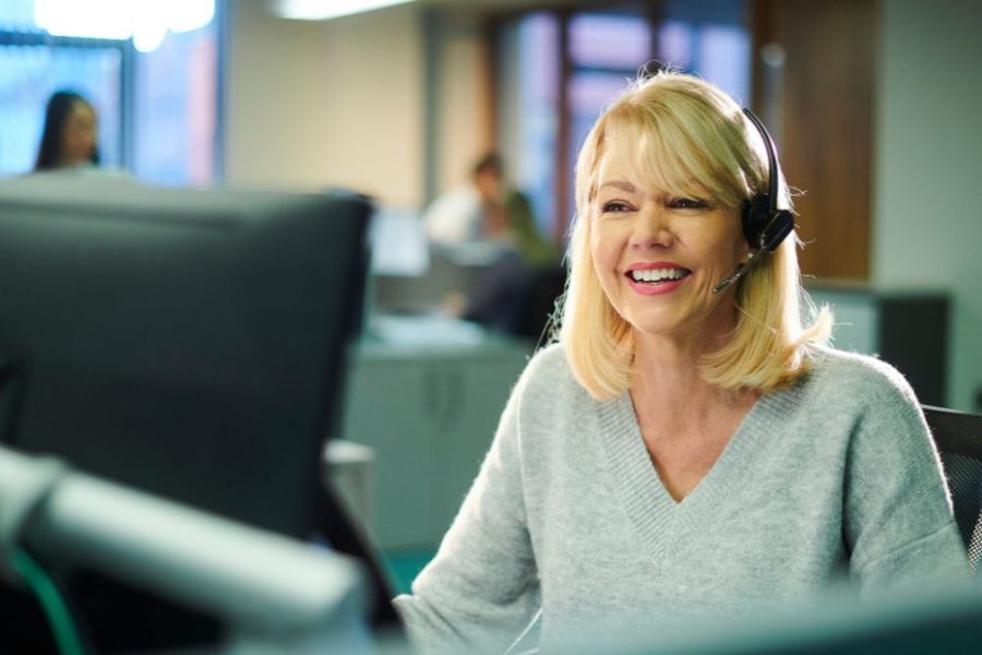 A member of staff, sat in front of her computer, smiling,  wearing a headset and talking to someone on the phone