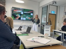 Someone delivering a presentation at the front of a room with attendees sitting around desks