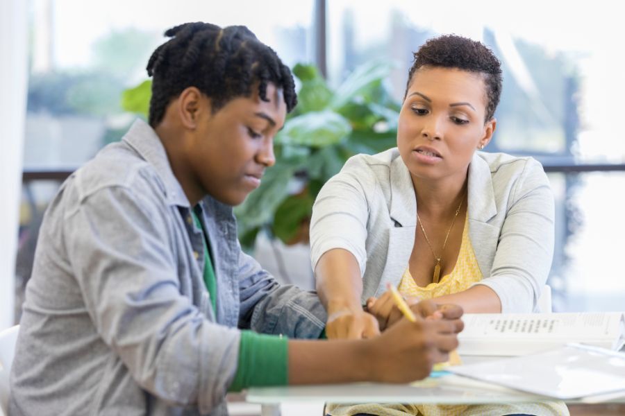 Teenager with mum at home both sat at a table looking at a school book.