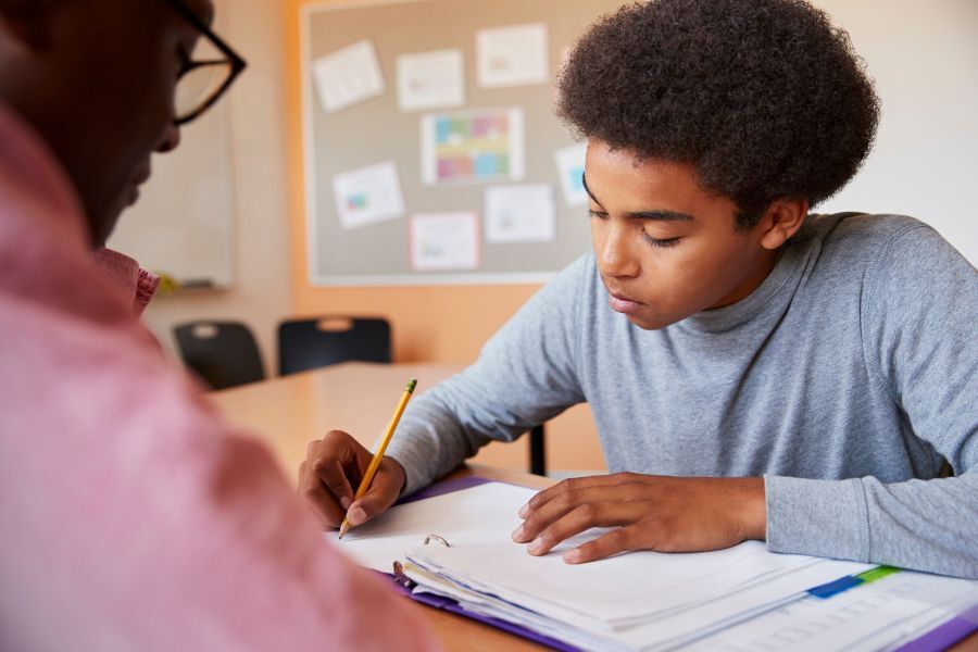 Boy sat with his teacher, one-to-one. Boy is writing in his book.