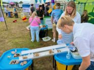 At the outdoor show stall, a boy and adult pour water into two sloped channels to compare flow over natural materials and an urban surface.