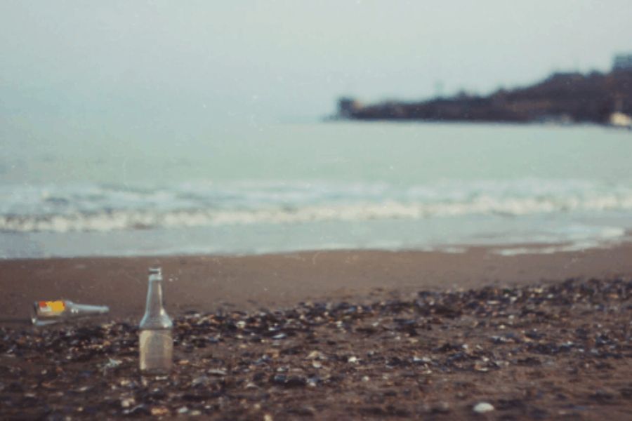 A pebbly beach, with the sea beyond. A beer bottle is stood upright on the pebbles