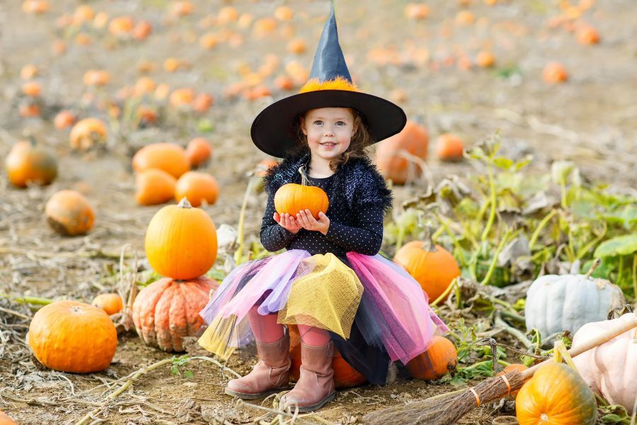 A young girl wearing a witch's hat and colourful skirt sat surrounded by pumpkins