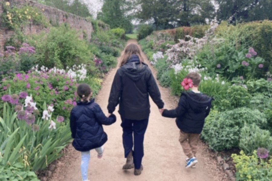 A mum and two children walking hand in hand through a garden, away from the camera