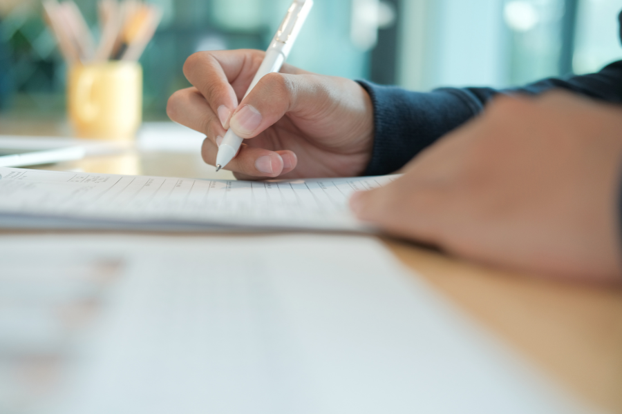 A close-up of a person writing on lined paper with a white pen, with additional sheets of paper and blurred stationery in the background on a desk