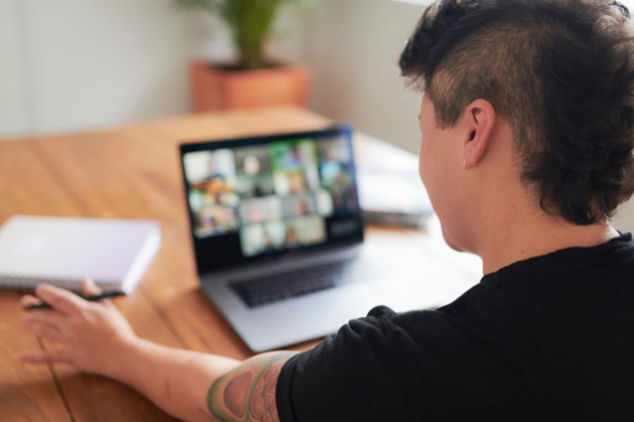 Person sitting at a wooden table using a laptop for a video conference. The laptop screen shows multiple participants in a grid layout.