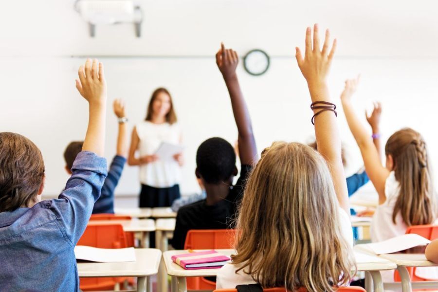 A classroom of pupils with their hands up to answer a question from the teacher at the front of the classroom