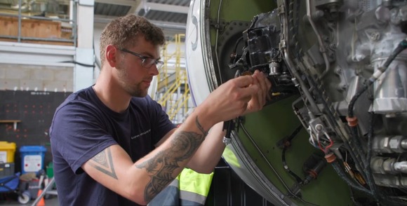 A person working on the internal components of an aircraft engine in an industrial workshop, wearing glasses and a dark blue shirt.