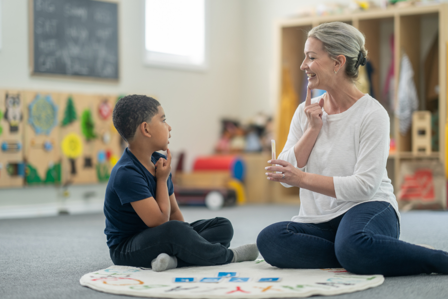 A speech and language therapist sitting on the floor with a child in a classroom, using a visual cue card during a communication exercise.