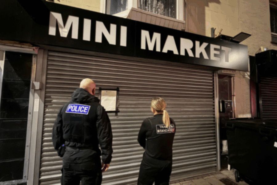 One police officer and one Trading Standards officer stood in front of a 'mini market' shop, that has its shutters down