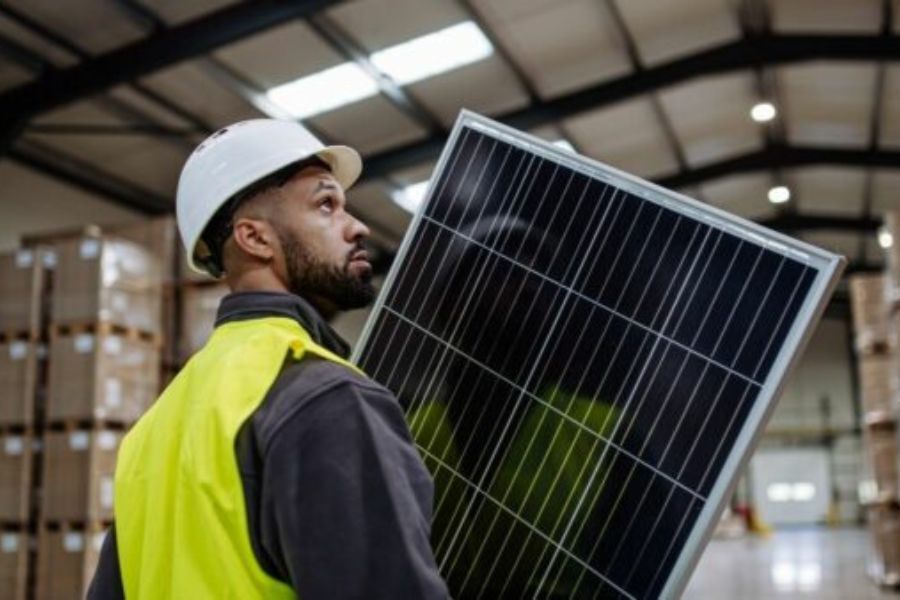 A worker in a hard hat and high-vis clothing, in a factory setting, carrying a solar panel
