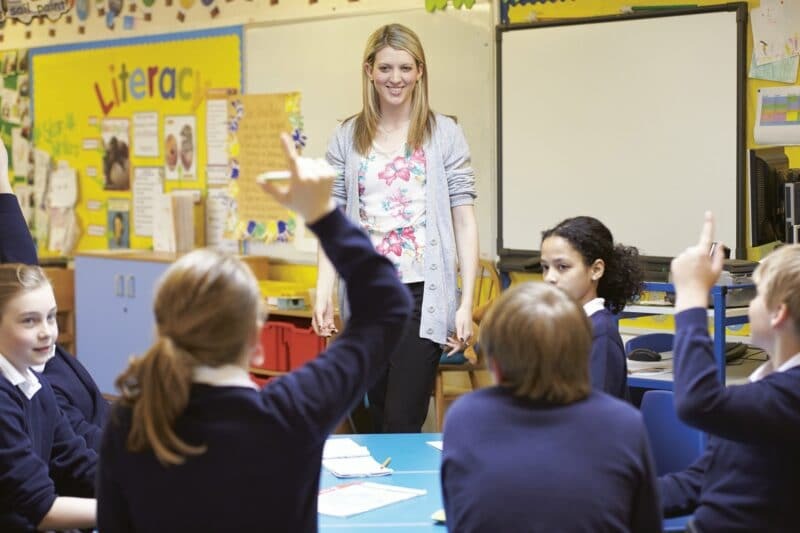 Students in a classroom raising hands while a teacher stands near a whiteboard and literacy display.
