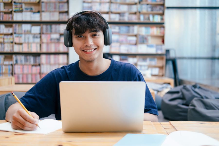 A college student, sat at a table in a library working, with head phones on