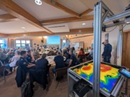 Interactive sandbox with contour lines projected onto its surface. In the background, adults are seated at tables.