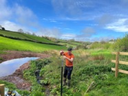 A man stands in a grassy field beside a large puddle, setting up a vertical measuring pole.
