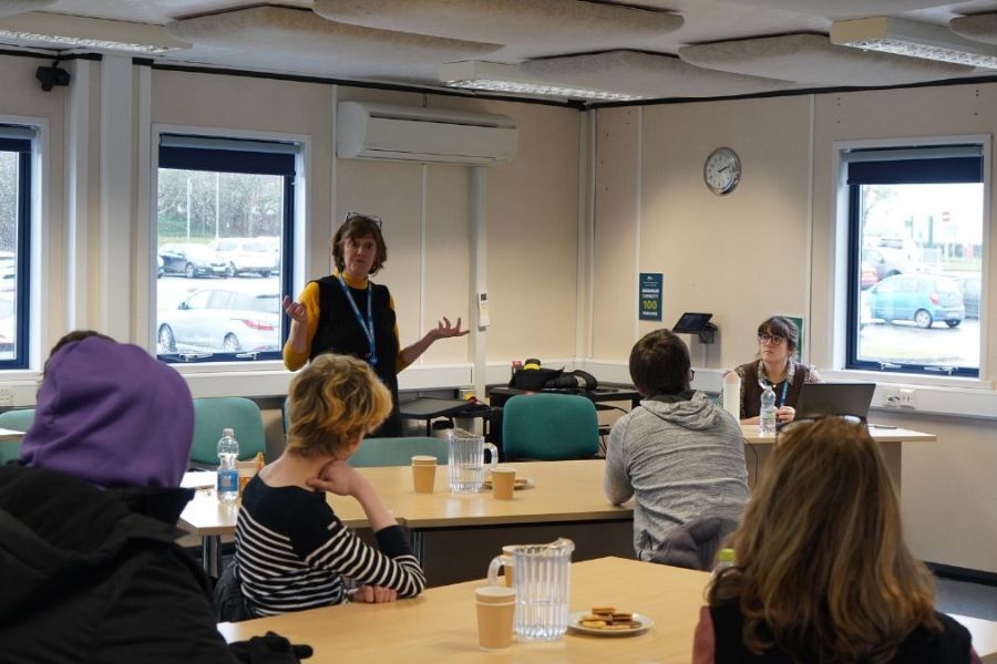 A group of people meeting in an office, sat around desks. There is a lead officer stood at the front of the room talking to the group