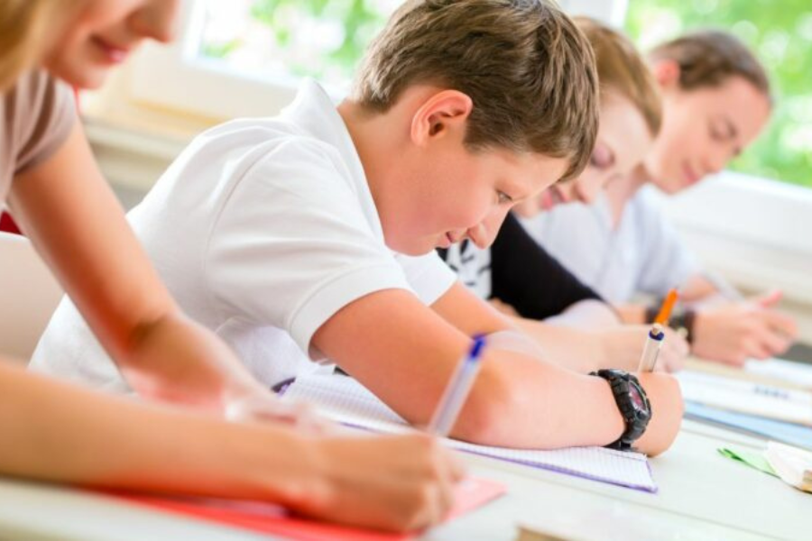 Children sit at desks in a classroom, focused on writing with pens and paper during a lesson.