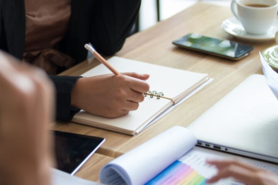 Adult taking notes in a notebook during a small meeting, with documents, a laptop and a phone on the table.