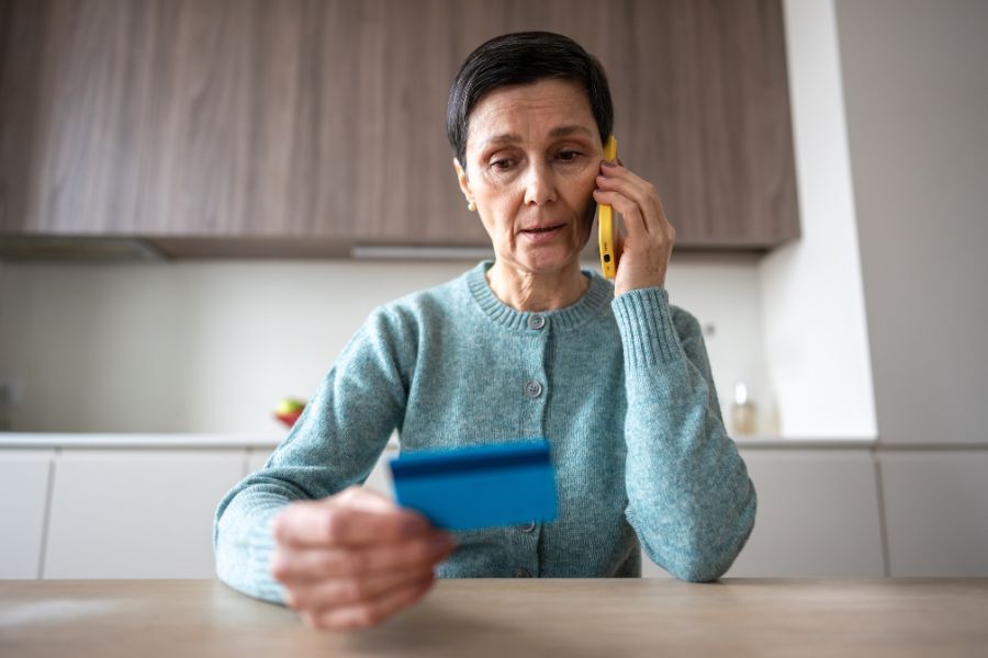 A lady sat at her kitchen table, on her mobile phone, looking at her credit card, as if making a payment.