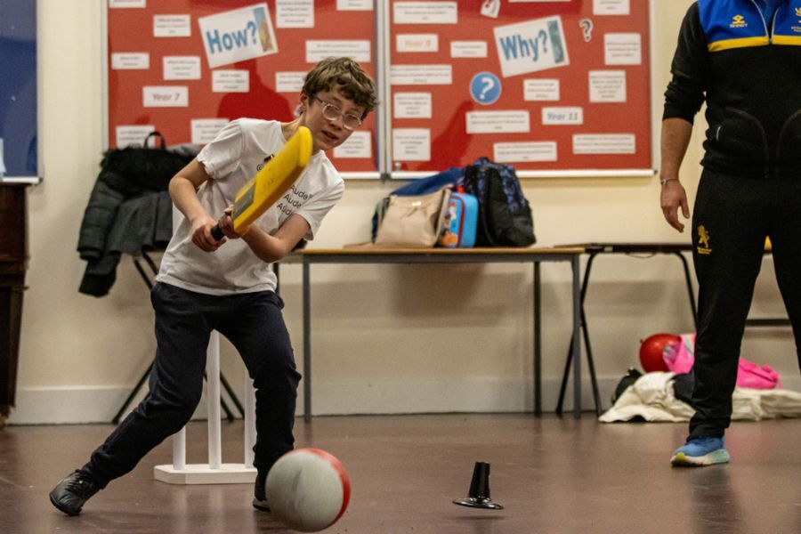A young boy playing with a cricket bat and football in a classroom, supervised with an adult