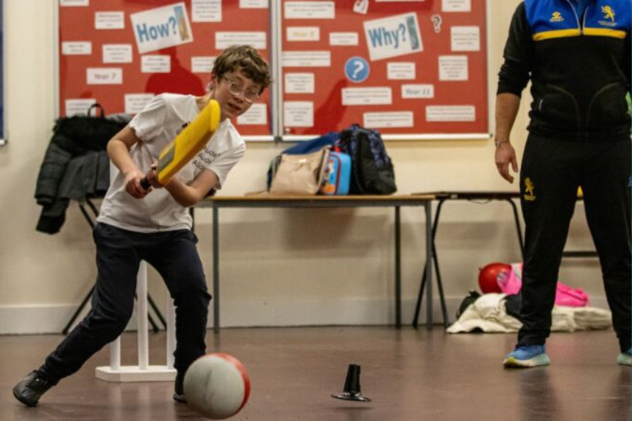 A child swings a plastic bat to hit a ball during an indoor sports activity, with an adult standing nearby.
