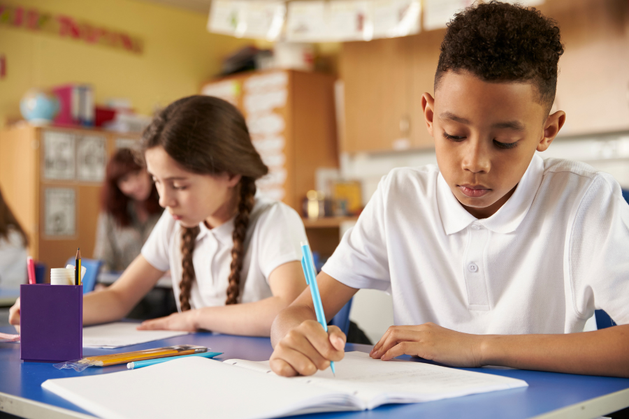 Two children sitting at a classroom table writing in their notebooks.