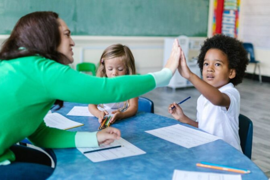 Adult and children sitting at a table doing schoolwork, with two giving a high‑five.