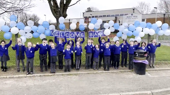 Children cheering outside standing underneath balloons and a sign reading 'welcome back'