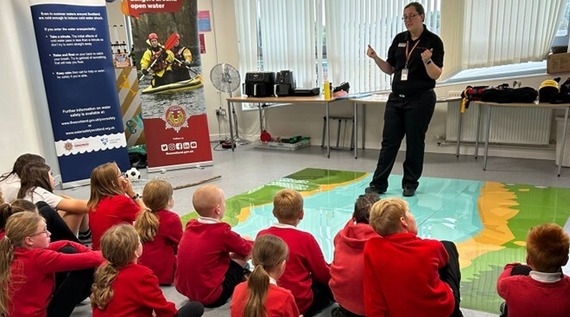 A picture depicts a police officer giving a talk to a group of primary school aged children sitting on the floor of a classroom