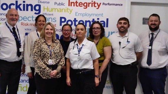 A picture depicts 8 mixed-sex staff members standing in front of a board showing empowering words. 