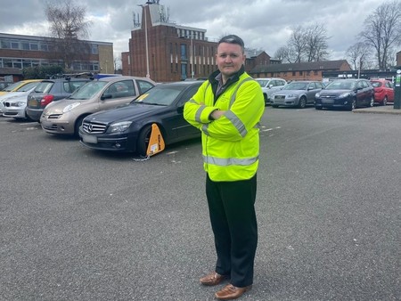 A man standing in front of a clamped vehicle in a car park.