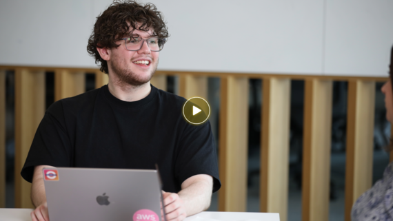Man smiling during a conversation at a laptop, with a blurred colleague and a gold play icon indicating video content.