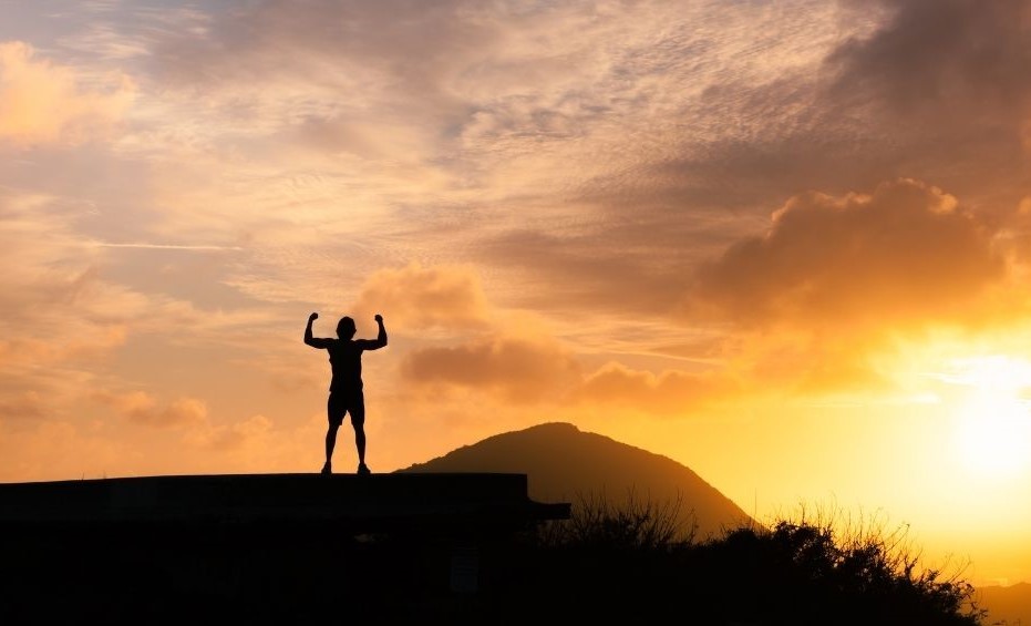 man looking triumphant at the top of a hill as the sun is setting