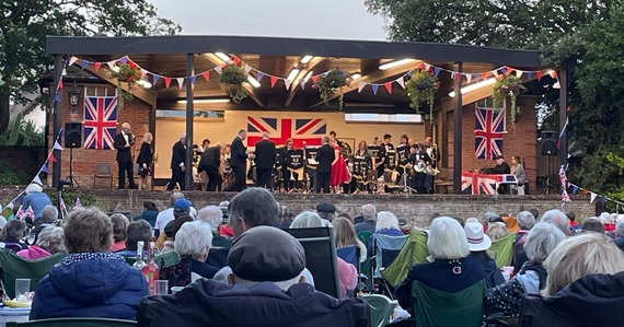 Sidmouth Town Band performing at Connaught Gardens. Union flags and bunting hang from the bandstand. The audience sits in front