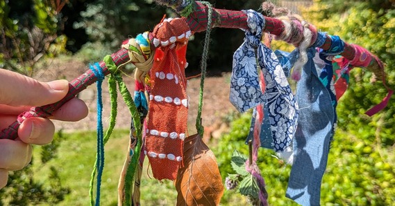 A hand holds a magic wand stick with fabric, leaves, and plants attached