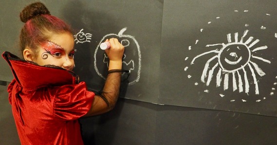 Child in fancy dress draws Halloween pictures in chalk on a large black piece of paper