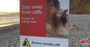 Sign post on a beach: Stay away from cliffs. Danger of falling rocks. Beware unstable cliff