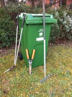 A green waste bin with gardening equipment propped against it