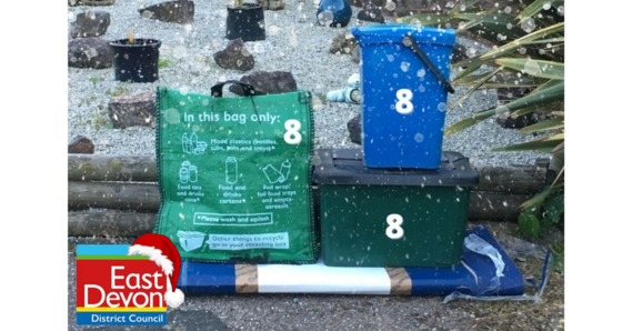 Snow falls on recycling containers. East Devon District Council logo with a Christmas hat
