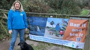 A person stands with their dog next to a 'bag it and bin it' banner that is attached to a countryside gate