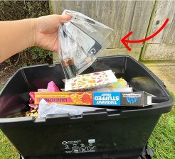 A hand holding a plastic bag with batteries in above a black recycling container with cardboard in it