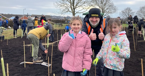 An adult and two children pose with thumbs up. In the background are volunteers planting trees at the tiny forest