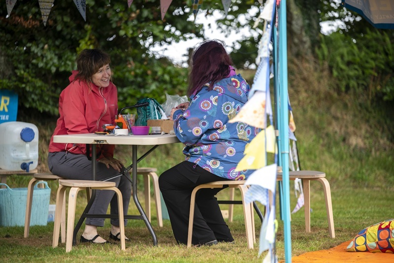 Two people sat opposite each other at at outside event. One person has a red hooded top on.