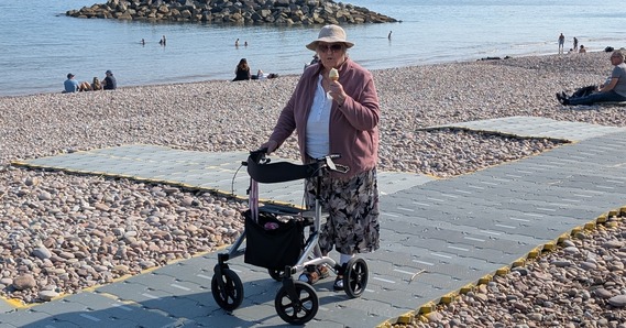 A person holds on to a stroller in one hand and an ice cream in the other. They are stood on matting on Sidmouth beach