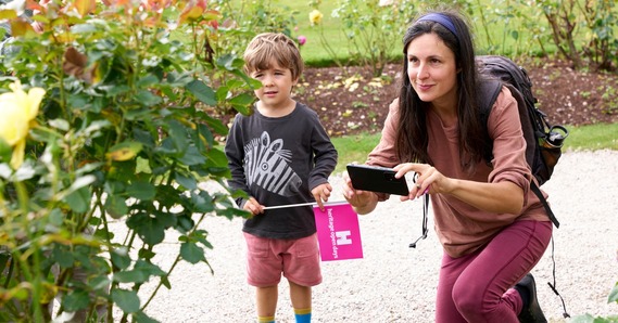 A child holding an Heritage Open Days flag stands next to a woman crouching to take a picture of a rose bush 