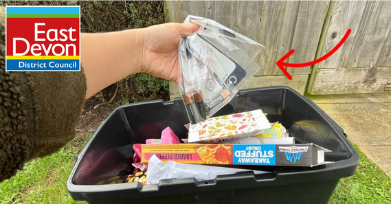 A hand holds a plastic bag of batteries above a recycling box