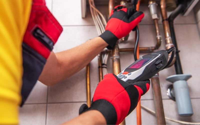 The hands of a boiler technician holding an electronic meter reader next to boiler pipes