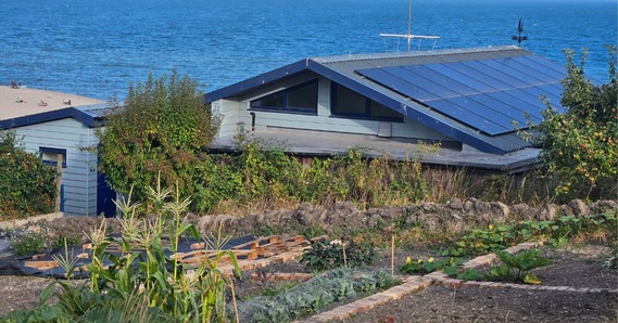 A building with solar panels on the roof, gardens in front, and the sea in the background