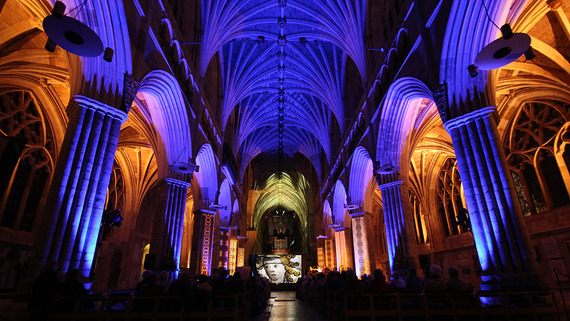 Blue lights illuminate the ceiling of Exeter Cathedral. An image of a person's face is in the background, behind rows of people on chairs